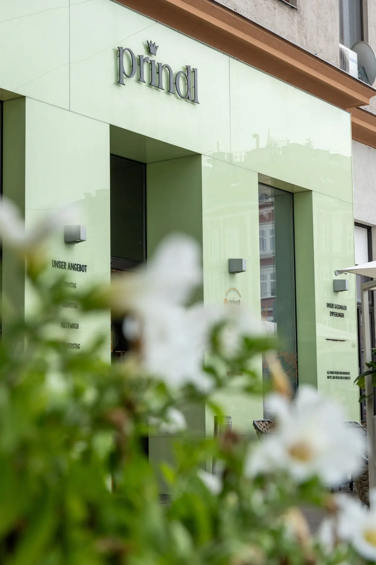 Prindl-Eingang mit grüner Fassade und Pflanzen im Vordergrund.