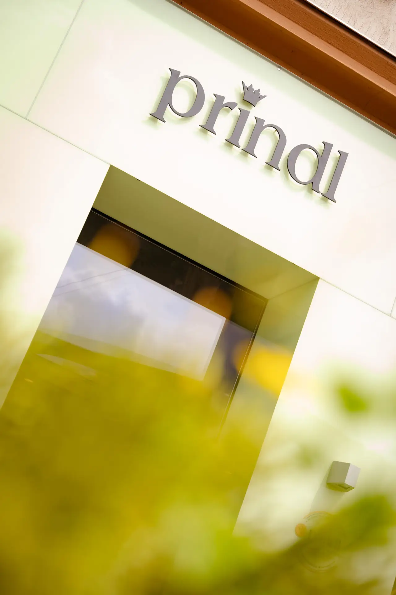 Prindl-Schriftzug über dem Eingang, durch unscharfe grüne Blätter fotografiert.